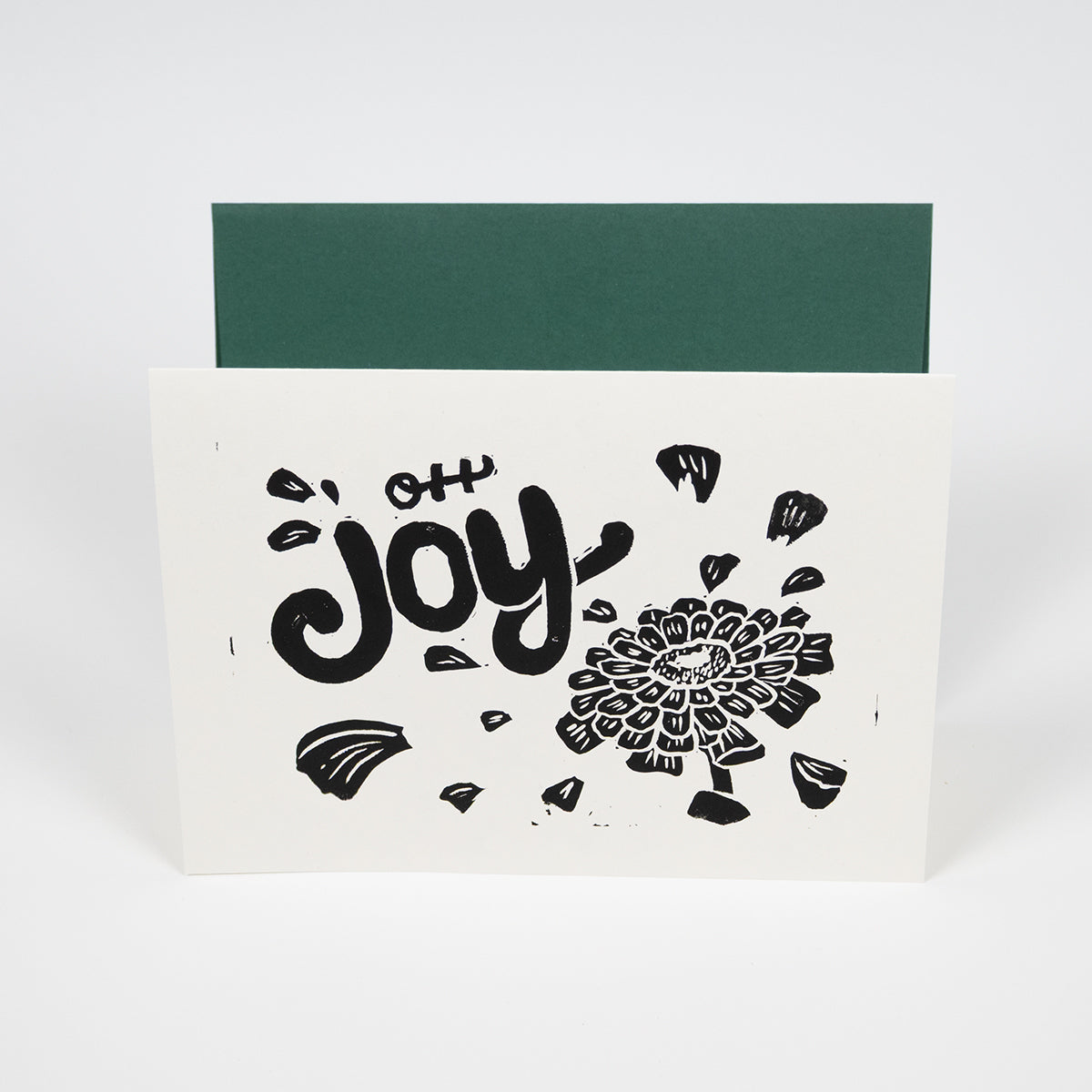 White greeting card with "Oh Joy" hand lettering with an illustration of a zinnia with petals floating around the text in front of a dark green envelope.
