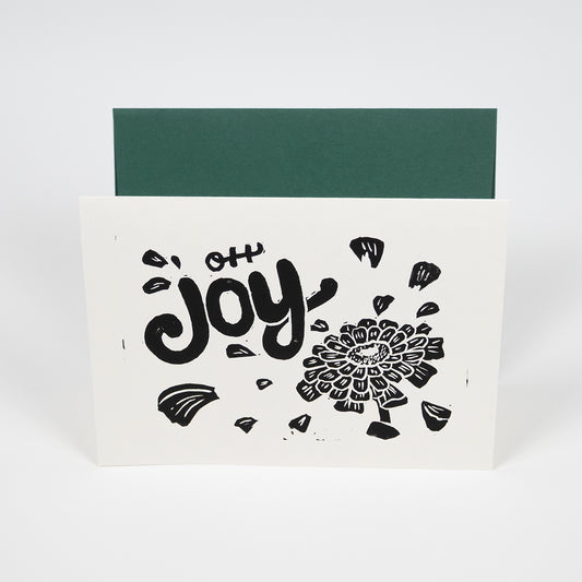 White greeting card with "Oh Joy" hand lettering with an illustration of a zinnia with petals floating around the text in front of a dark green envelope.