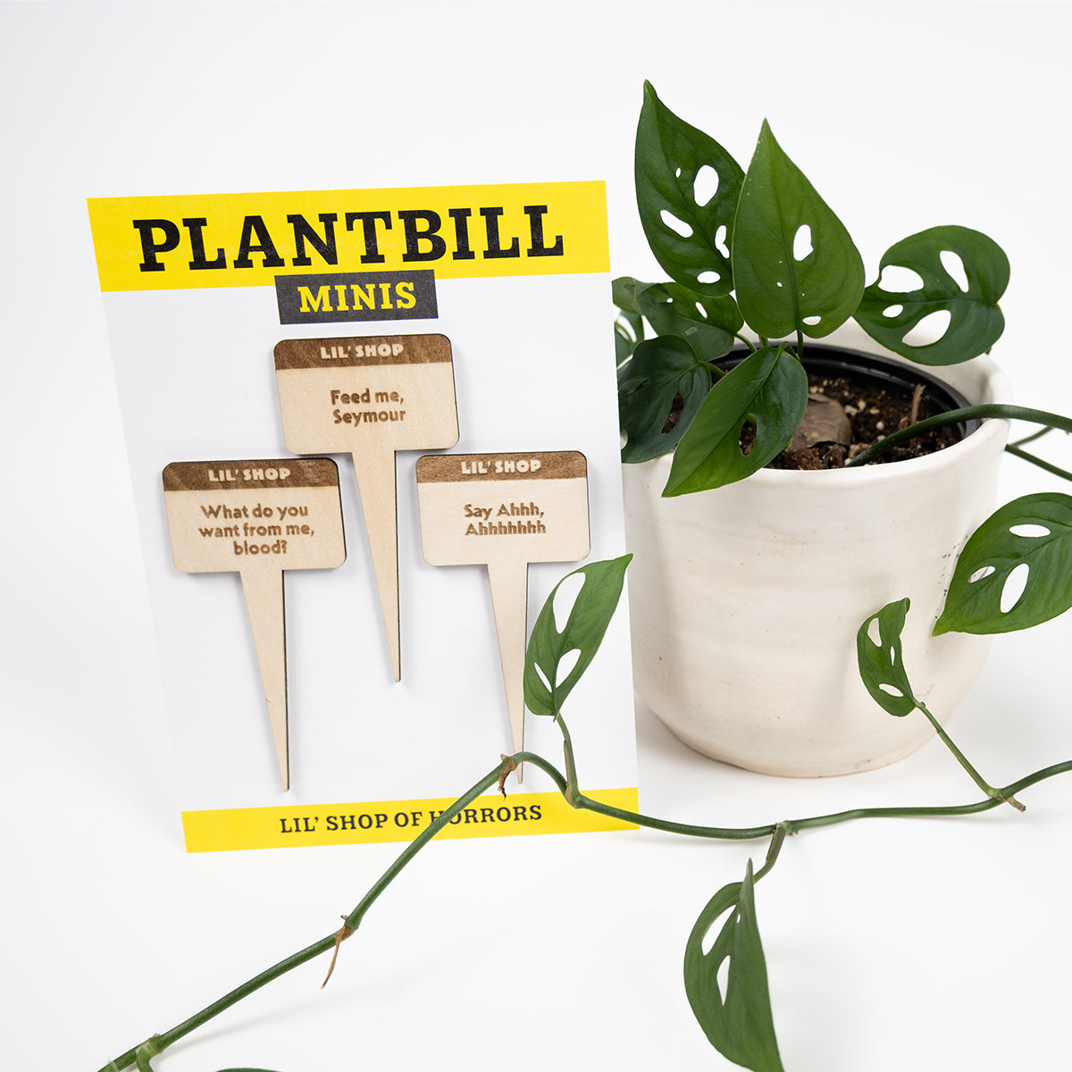 White and yellow card with "Plantbill" across the top and "Lil' Shop of Horrors" across the bottom. In the middle are two wooden plant/garden stakes with "What do you want from me, blood?," "Feed me, Seymour," and "Sa Ahh, Ahhhhh" engraved on them paired with a swiss cheese plant in a white planter.
