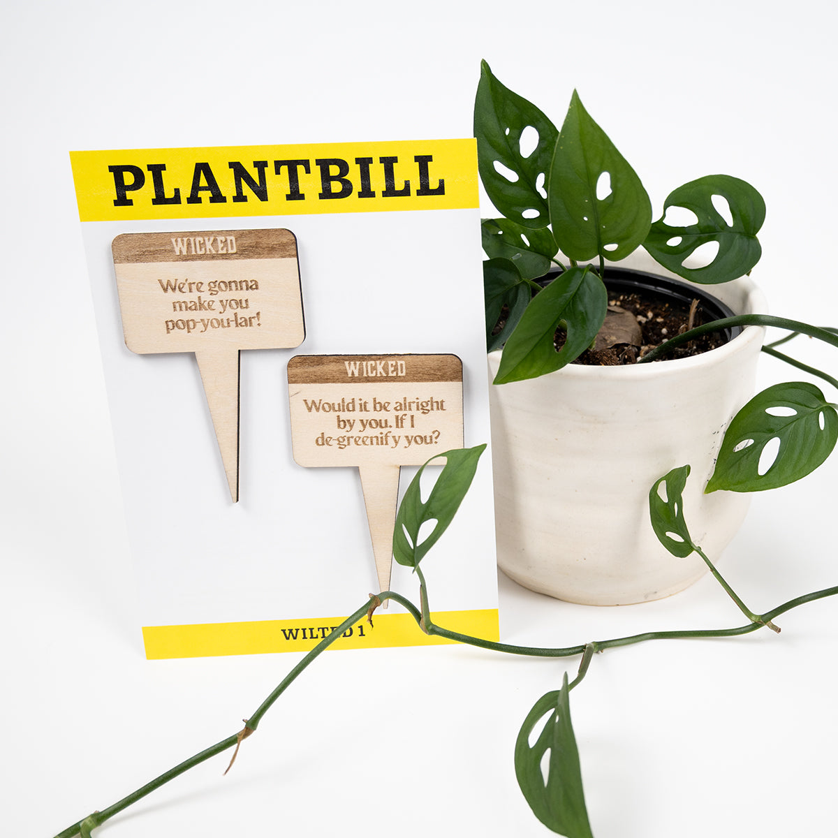 White and yellow card with "Plantbill" across the top and "Wilted 1" across the bottom. In the middle are two wooden plant/garden stakes with "We're gonna make you pop-you-lar" and "Would it be alright by you. If I de-greenify you?" engraved on them paired with a swiss cheese plant in a white planter.