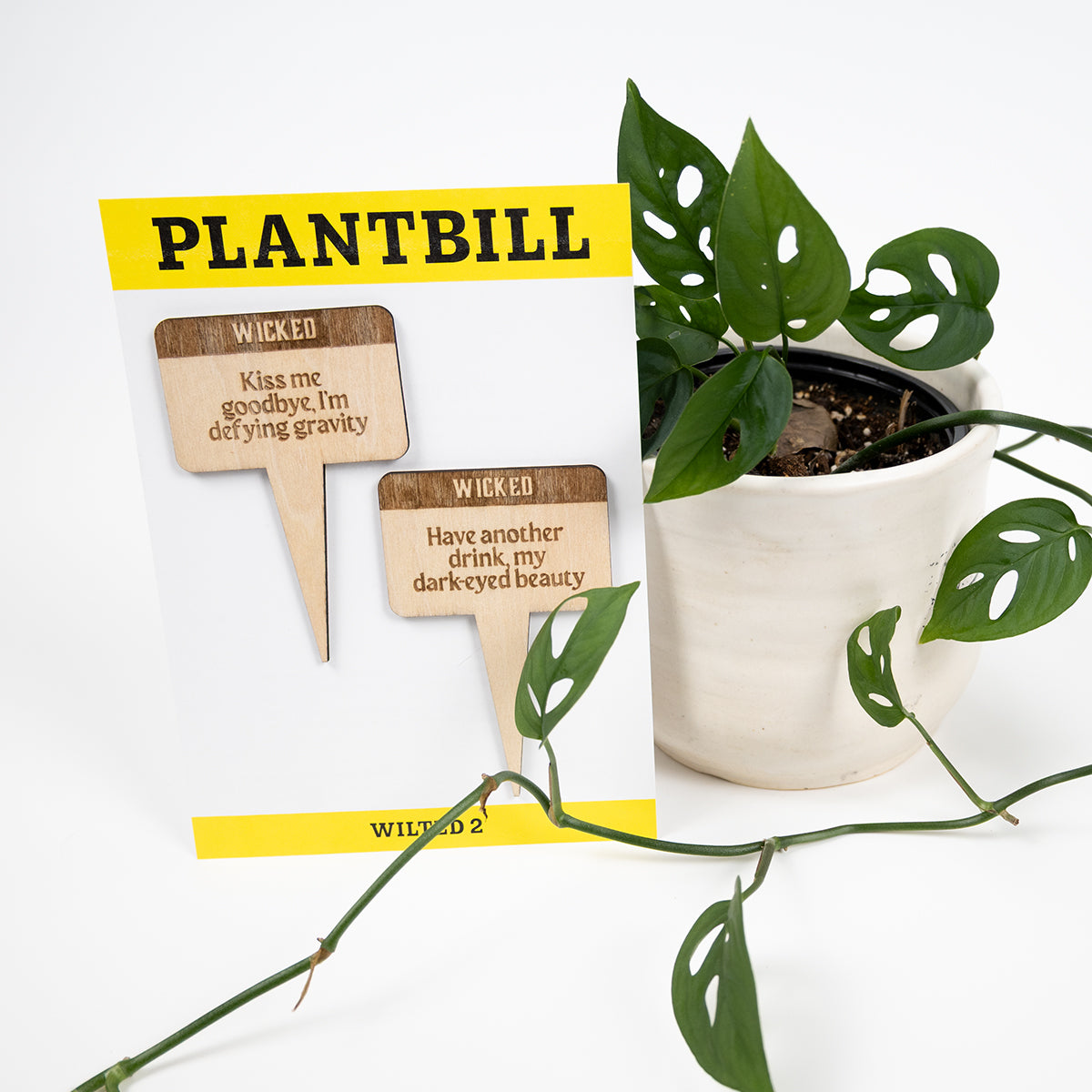 White and yellow card with "Plantbill" across the top and "Wilted 2" across the bottom. In the middle are two wooden plant/garden stakes with "Kiss me goodbye, I'm defying gravity" and "Have another drink, my dark-eyed beauty" engraved on them paired with a swiss cheese plant in a white planter.