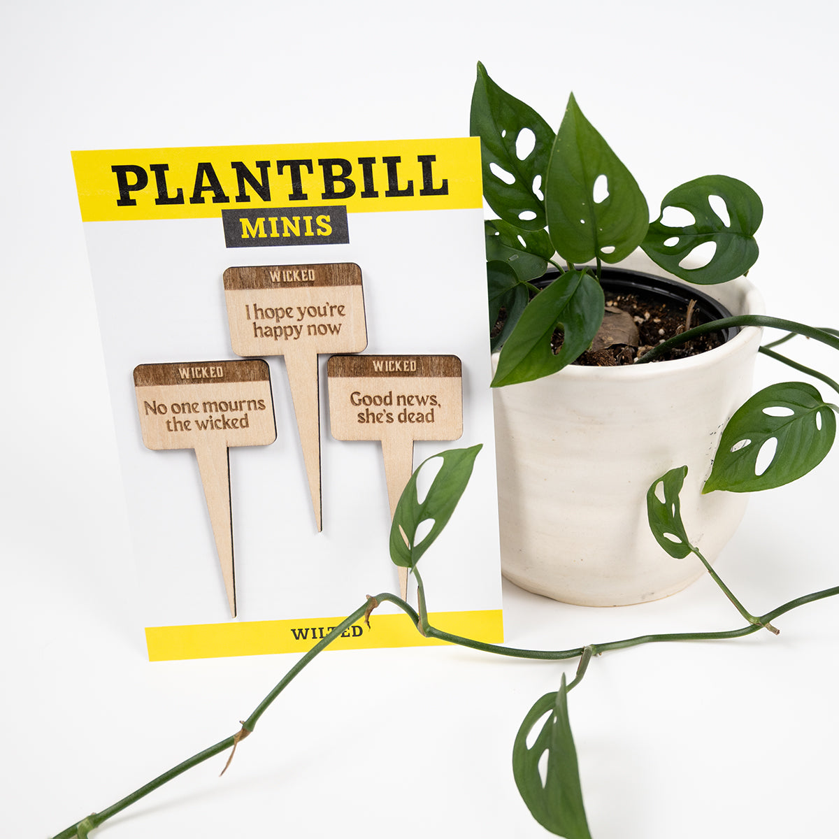 White and yellow card with "Plantbill Minis" across the top and "Wilted" across the bottom. In the middle are three wooden plant/garden stakes with "No one mourns the wicked," "I hope you're happy now," and "good news, she's dead" engraved on them paired with a swiss cheese plant in a white planter.