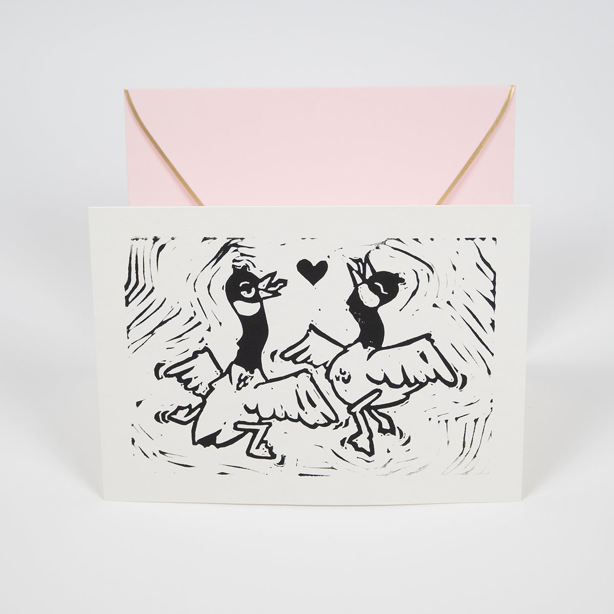 White greeting card with illustration of two silly, dancing geese with a heart between them in front of a light pink envelope.