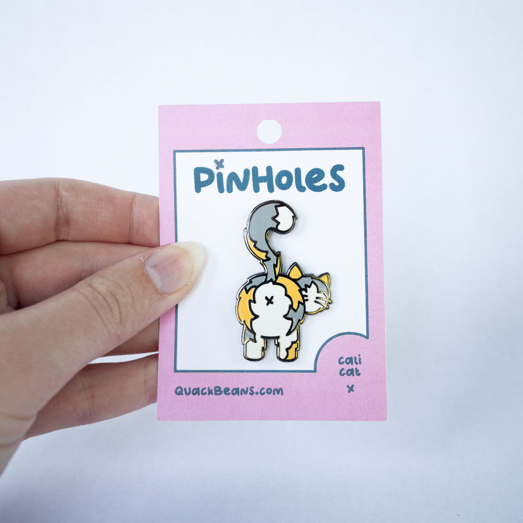 Hand holding pink Pinholes hang tag card with a light calico cat butt pin.