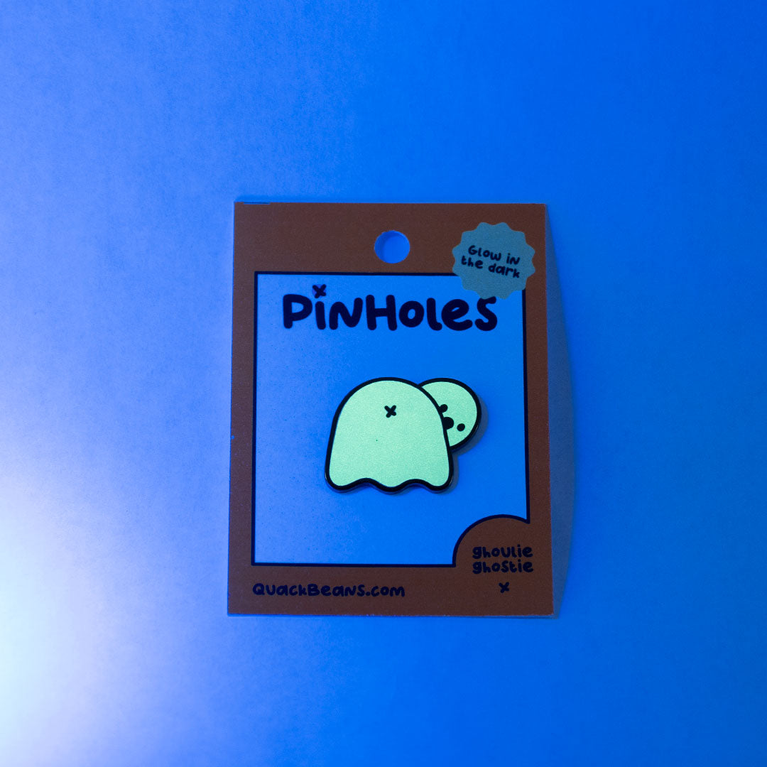 Dark view of a glow in the dark ghost butt pin on a orange backing card with the word "Pinholes"