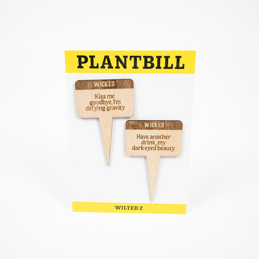 White and yellow card with "Plantbill" across the top and "Wilted 2" across the bottom. In the middle are two wooden plant/garden stakes with "Kiss me goodbye, I'm defying gravity" and "Have another drink, my dark-eyed beauty" engraved on them. 