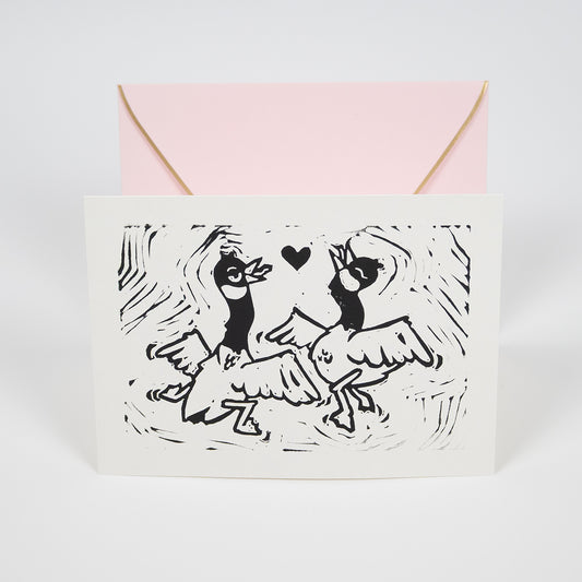 White greeting card with illustration of two silly, dancing geese with a heart between them in front of a light pink envelope.